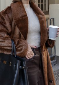 Manteau en fausse fourrure marron avec larges revers, pull crème, jupe en cuir marron, sac à main noir et tasse de café à emporter à la main.
