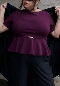 Haut péplum violet à manches courtes, taille cintrée et détail de boucle de ceinture argentée. Tissu noir drapé tenu dans les mains.