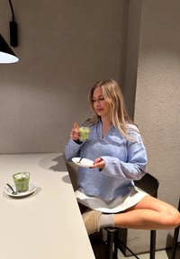 Ανοιχτό μπλε, oversized πουλόβερ με γιακά; λευκή φούστα. Κρατάει ένα πράσινο ποτό σε ποτήρι και ένα λευκό πιάτο με κουτάλι. Ουδέτερο φόντο.