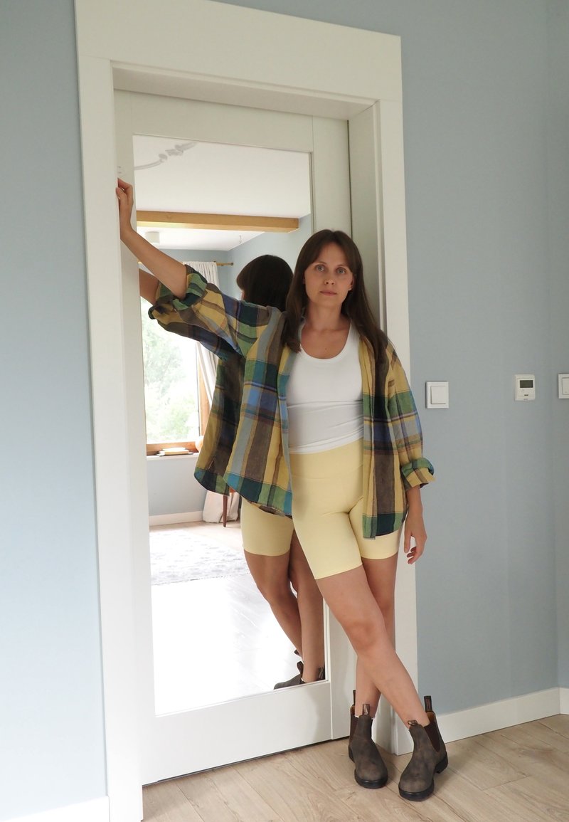 Femme en chemise à carreaux, débardeur blanc, short jaune et bottes marron appuyée contre un cadre de porte blanc avec un miroir derrière elle.