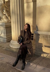 Manteau marron porté sur un pull sombre, avec une jupe marron fluide et des bottes noires montantes. Tenant un sac à main noir, assise près de colonnes en pierre.