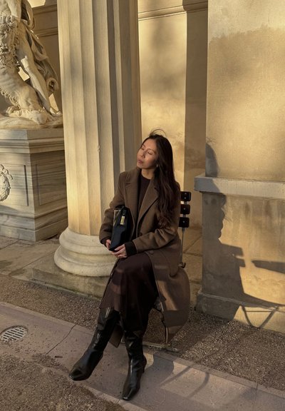 Abrigo marrón sobre un suéter oscuro, combinado con una falda marrón fluida y botas negras hasta la rodilla. Sosteniendo un bolso negro, sentada junto a columnas de piedra.