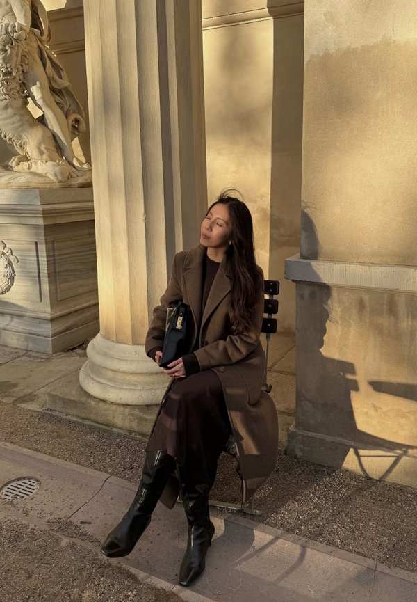 Brun överrock över en mörk tröja, kombinerad med en flödande brun kjol och knähöga svarta stövlar. Håller en svart handväska, sittande vid stenkolonner.