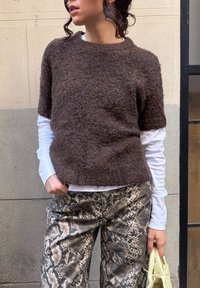 Femme portant un pull court à manches courtes marron duveteux sur une chemise blanche à manches longues, un pantalon imprimé serpent, et tenant un petit sac à main jaune pâle.