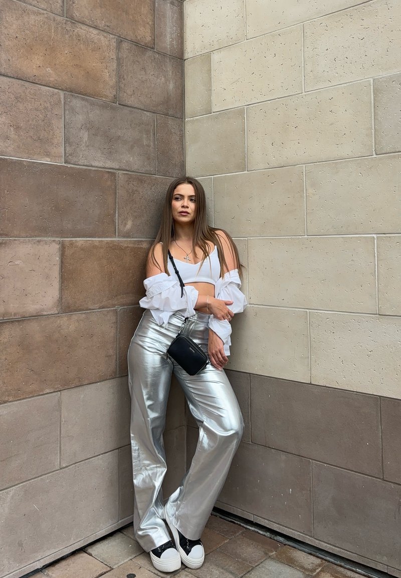 Sølv, vide metalliske bukser, hvit kortermet topp og lagdelte gjennomsiktige hvite ermer. Svart crossover-veske og joggesko. Teksturrik vegg bakgrunn.
