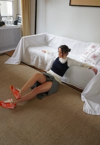 Una persona está sentada sobre una alfombra beige frente a un sofá blanco, llevando una camiseta oscura sin mangas, una falda gris y zapatillas Adidas de color naranja brillante.