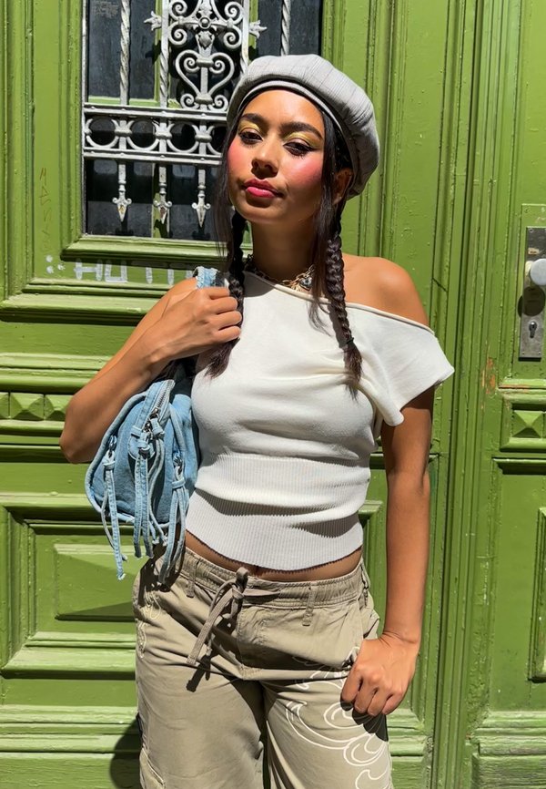 Ung kvinde med flettet hår iført en grå baret og hvid off-shoulder top, stående foran en grøn udsmykket dør med en lyseblå taske i hånden.
