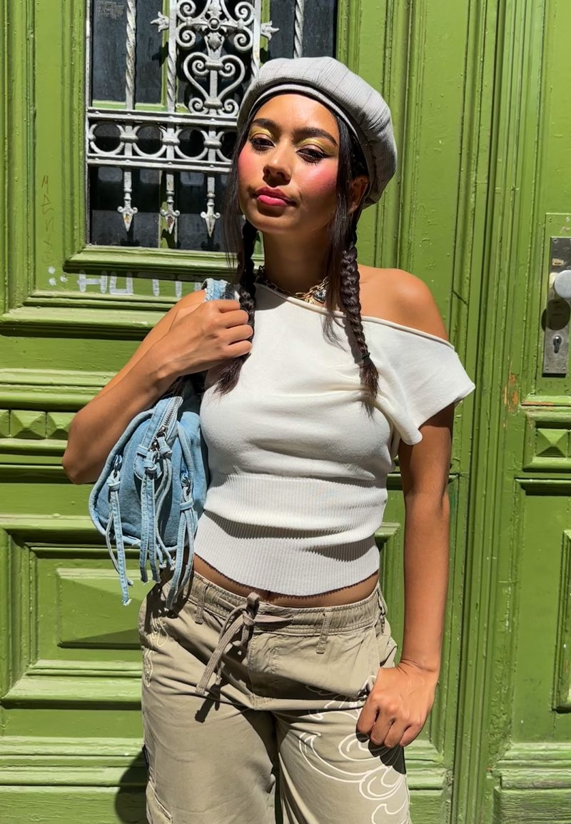 Jeune femme aux cheveux tressés portant un béret gris et un haut blanc épaules dénudées, debout devant une porte verte ornée tenant un sac bleu clair.
