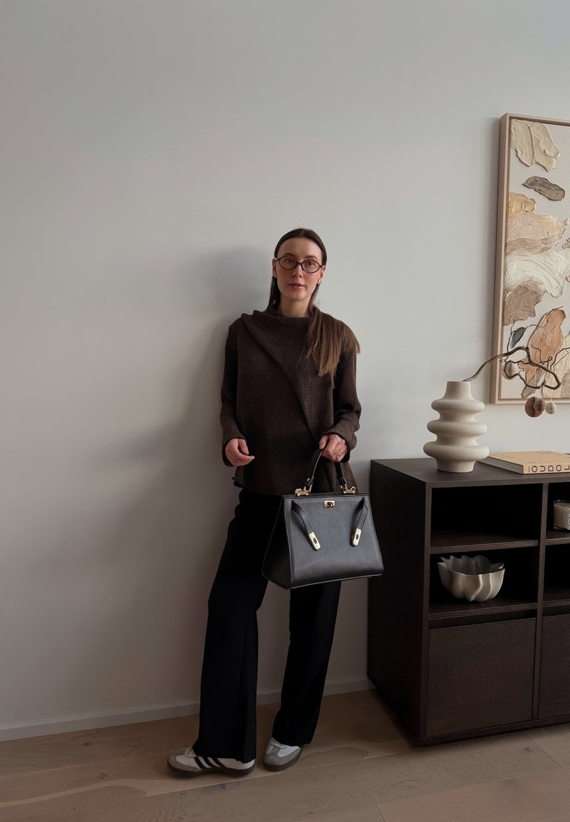 Femme portant des lunettes, un pull marron foncé et un pantalon noir, tenant un sac à main noir, debout à côté d'une armoire en bois foncé avec décoration.
