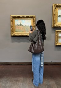 Ruskea punottu nahkalaukku löysällä mallilla, yhdistettynä väljiin sinisiin farkkuihin ja paksuun harmaaseen neuleeseen, nähtynä taidegallerian ympäristössä.