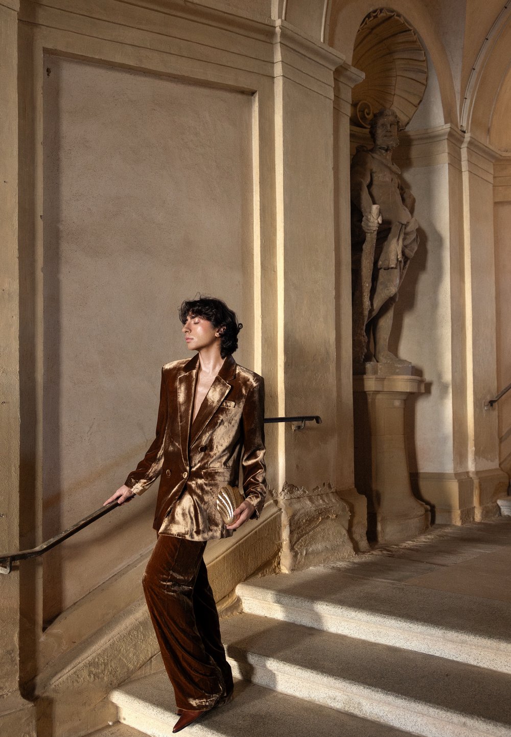 Personne en costume en velours brun brillant et talons descendant un escalier en pierre à l'intérieur d'un bâtiment historique avec une statue classique sur le mur.