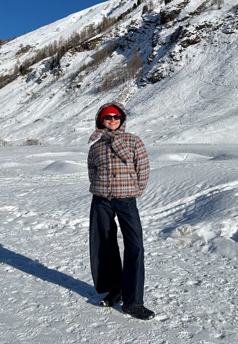 Personne portant une doudoune à carreaux, un bonnet rouge, des lunettes de soleil et un pantalon noir, debout sur un sol enneigé avec une montagne couverte de neige en arrière-plan.
