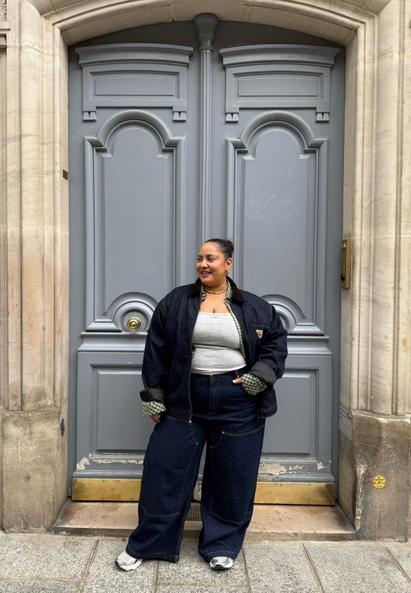 Femme avec un chignon foncé, portant une veste en jean foncé et un pantalon avec un haut gris, debout en souriant devant de grandes portes doubles grises ornées sur un bâtiment en pierre.