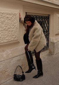 Túlméretezett bézs bunda, térd fölé érő fekete fényes csizma, földön fekvő fekete táska, textúrált kőfal mellett áll.
