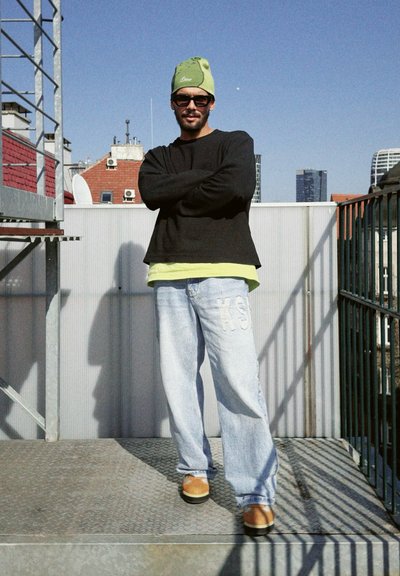 Camisa negra de mangas largas sobre una camiseta verde lima, jeans azul claro con bordado "KS", gorro verde y zapatos marrones, de pie en una azotea.