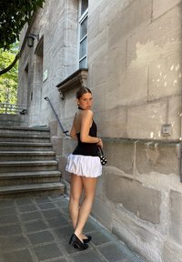 Top negro sin mangas combinado con una falda corta blanca con volantes. Zapatos de tacón negros y un bolso de lunares. El fondo presenta una pared de piedra y escaleras.