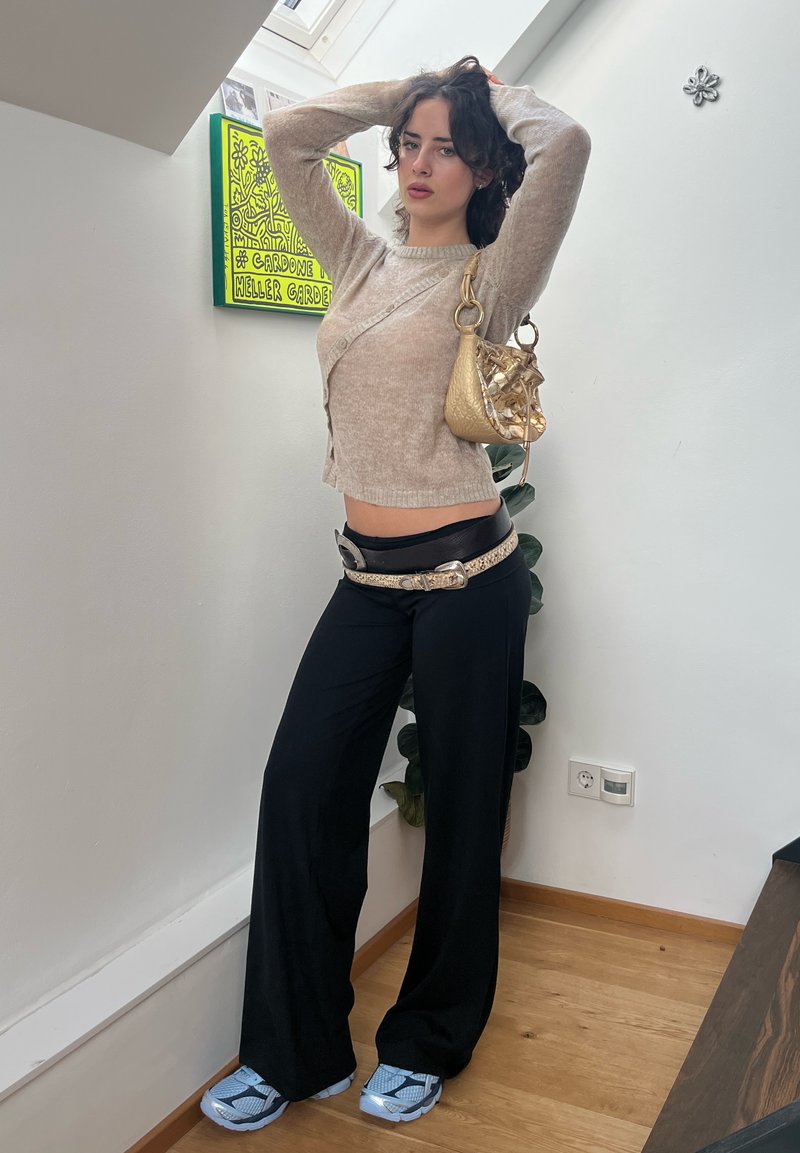 Šviesiai smėlinė megztinė, juodos plačios kelnės su dekoratyvine diržu ir mėlyni sportbačiai. Laiko auksinę rankinę. Patalpa su augalais.