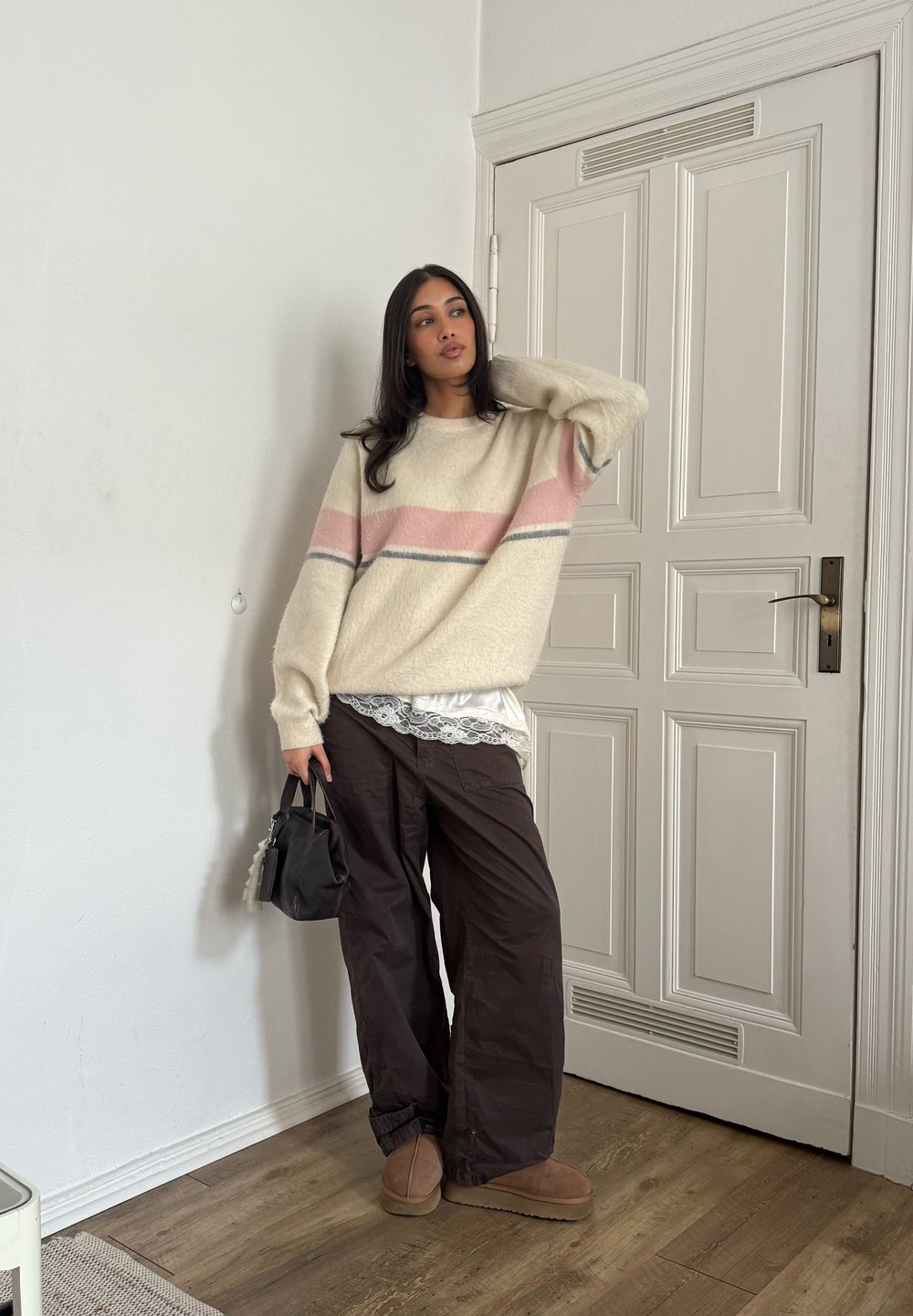 Pull-over blanc cassé avec des rayures roses et grises, assorti à un pantalon ample marron foncé. Accessoire : un petit sac noir. Texture lisse.