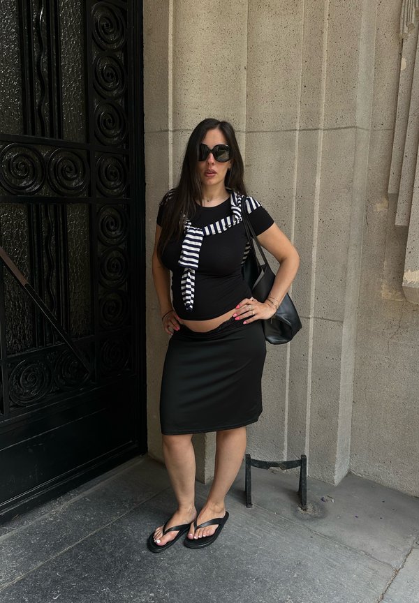 Femme aux longs cheveux foncés et lunettes de soleil, debout avec une main sur la hanche, portant un haut noir, une jupe, un pull rayé et des tongs près d'un mur en pierre.