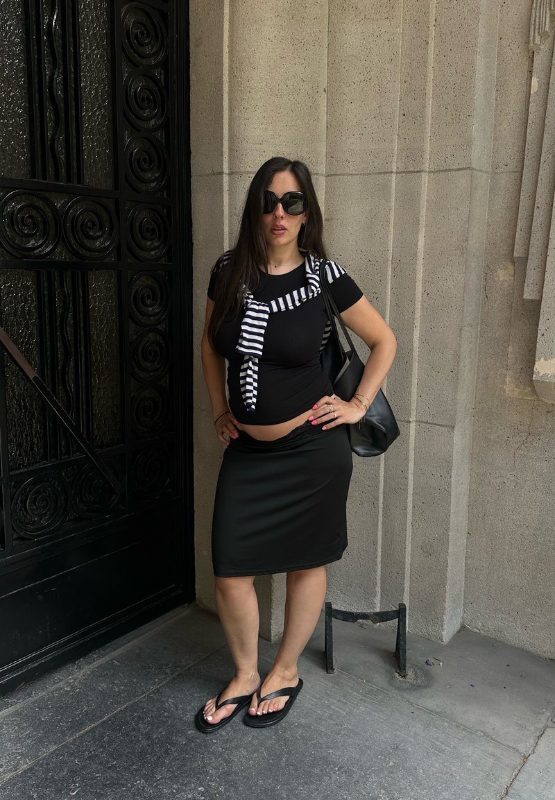Femme aux longs cheveux foncés et lunettes de soleil, debout avec une main sur la hanche, portant un haut noir, une jupe, un pull rayé et des tongs près d'un mur en pierre.