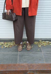 Personne portant un pantalon large marron, des chaussures à imprimé léopard et une veste en velours côtelé orange, tenant un sac à main marron foncé, debout sur un sol carrelé parsemé de feuilles.