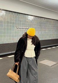 Person wearing a yellow beanie, black coat, white blouse, and grey trousers stands on a subway platform holding a beige handbag, looking down.