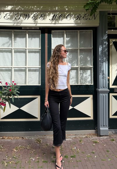 Una mujer se encuentra junto a un edificio, vistiendo una blusa blanca ajustada y pantalones negros recortados con borde de encaje, mientras sostiene un bolso negro.