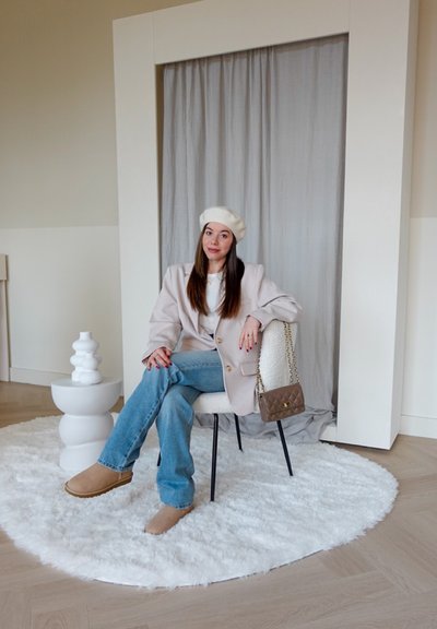 Una persona sentada en un blazer rosa claro de gran tamaño y una boina blanca, vestida con jeans azules y zapatillas beige sobre una alfombra redonda y esponjosa junto a una mesa escultórica blanca.