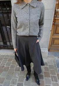 Graue Wollstrickjacke mit Kragen und Knopfverschluss, kombiniert mit einem dunkelgrauen knielangen Rock. Schwarze kniehohe Stiefel mit spitzen Zehen und eine kleine schwarze Tasche.