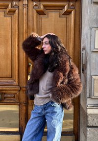 Femme aux longs cheveux foncés, portant une veste en fourrure marron et un pull beige, debout devant une porte en bois ornée, main sur la tête, regardant sur le côté.
