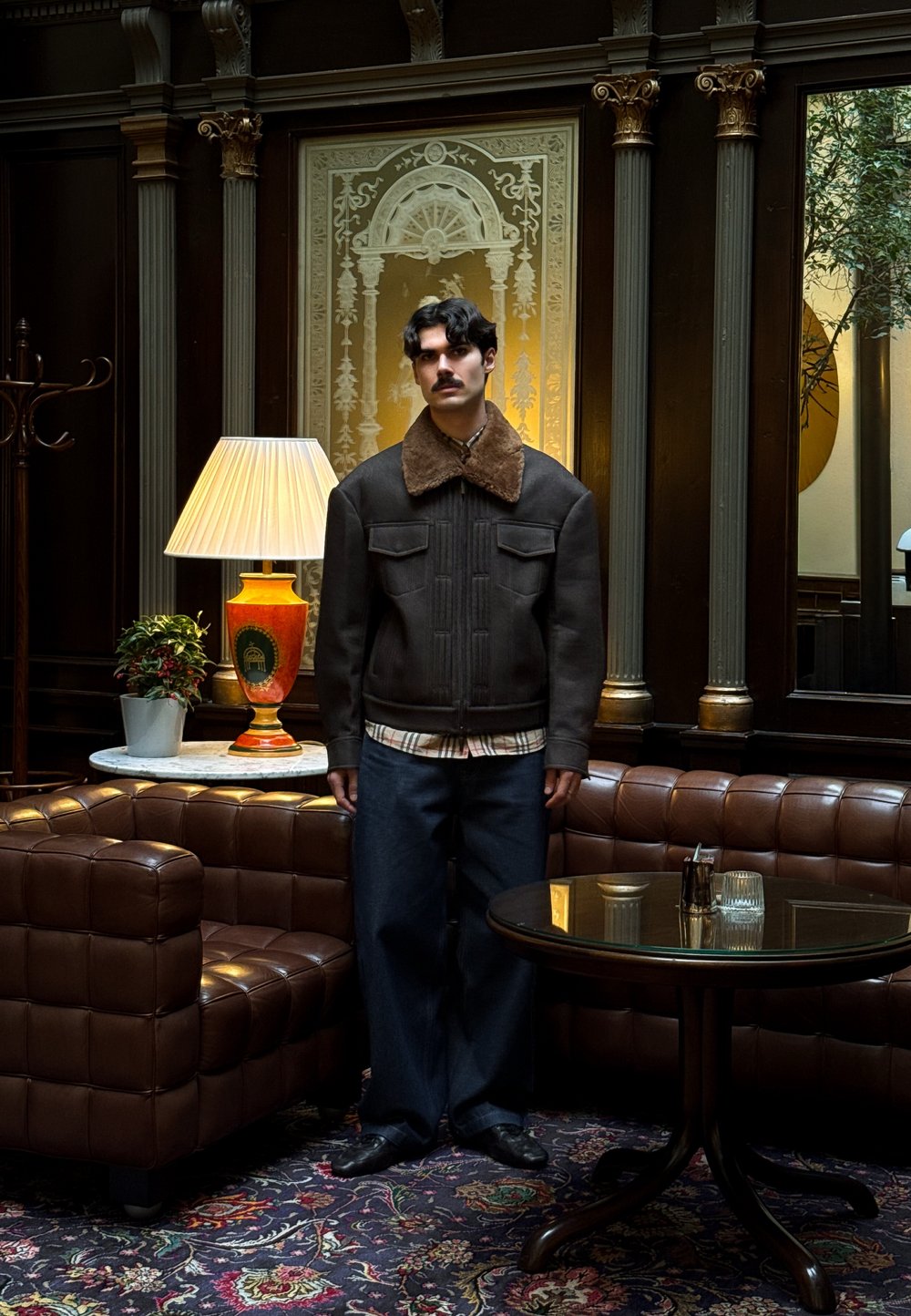 Veste marron à col en peau retournée avec poches avant, associée à un pantalon en denim foncé. Un homme se tient près d'un canapé en cuir marron et d'une lampe.