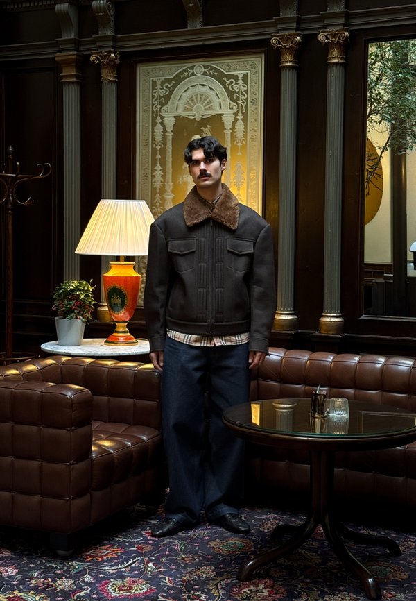 Rjava jakna s podlogo iz ovčjega kožuha in žepi spredaj, kombinirana z temnimi denim hlačami. Moški stoji ob rjavi usnjeni sedežni garnituri in svetilki.