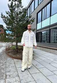 Krämfärgad linneskjorta med blå broderidetaljer, i kombination med lösa krämfärgade cargobyxor och vita sneakers, placerad i en urbant miljö.