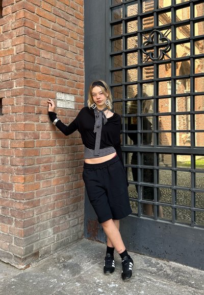 Top negro de manga larga con una bufanda a rayas, pantalones cortos negros y zapatillas negras. Una pared de ladrillo y una ventana enrejada proporcionan un fondo texturizado.