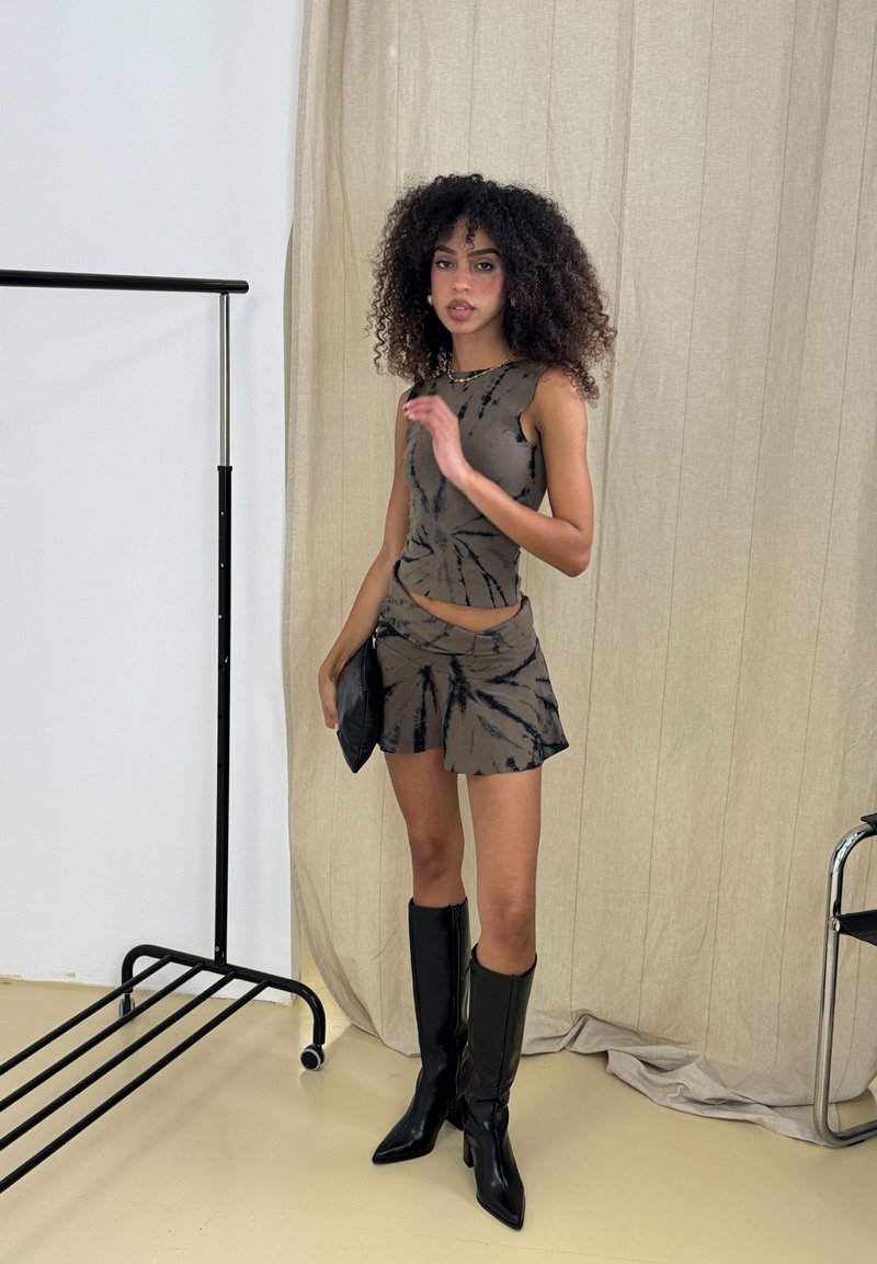 Conjunto de duas peças em tie-dye castanho com top cropped sem mangas e saia mini. A modelo usa botas de salto alto pretas até ao joelho e segura uma clutch preta.