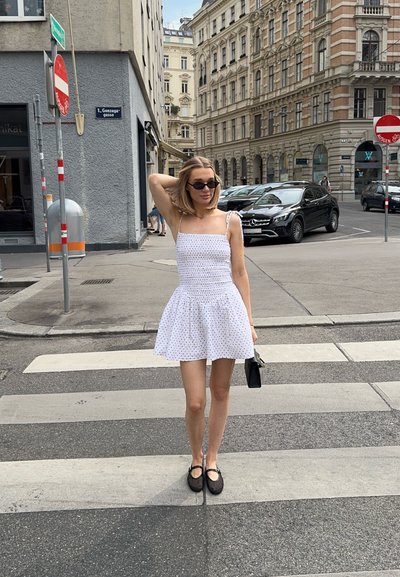 Vestido blanco con lunares negros, falda corta, tirantes delgados. Bolso negro y zapatos. Entorno urbano con paso de peatones y edificios.