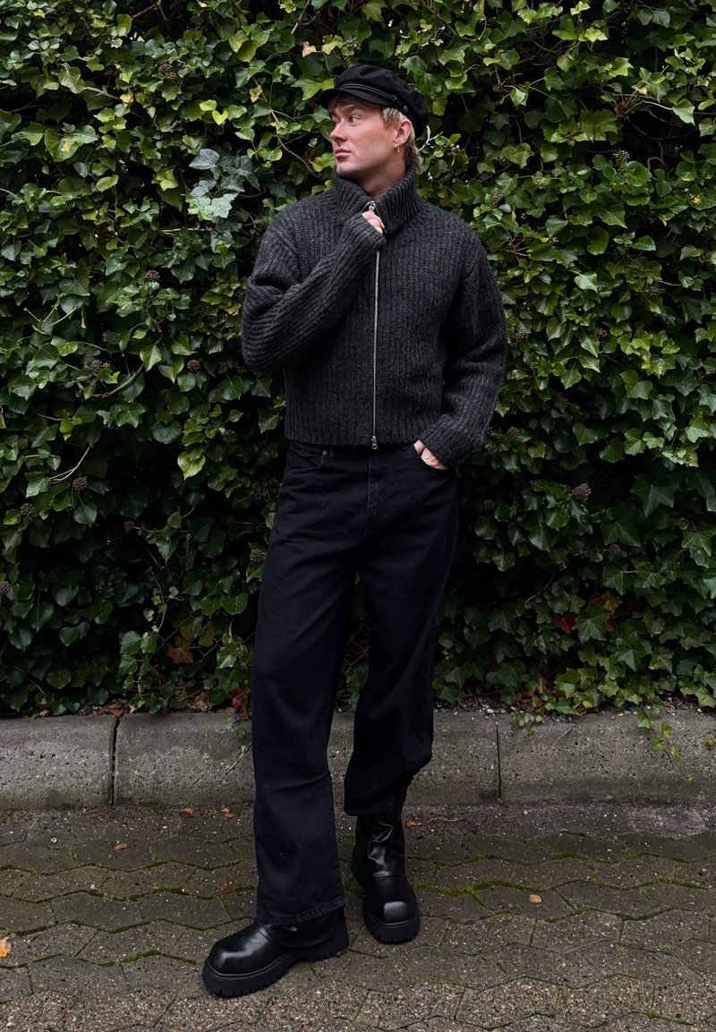 Maglione nero a coste con colletto alto e chiusura con zip. Abbinato a pantaloni neri a gamba larga e stivali neri robusti. Sfondo di edera verde.