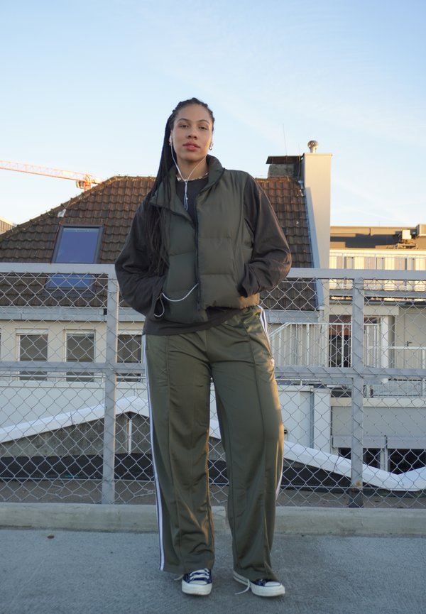 Grønn puffervest med svarte ermer, parret med olivengrønne bukser og marineblå sneakers. Øretelefoner på, stående mot en takbakgrunn.