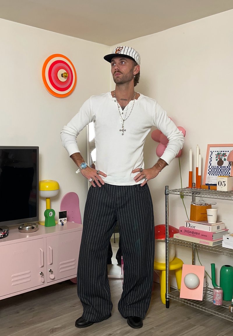 Camicia bianca a maniche lunghe, pantaloni larghi a righe e un cappellino da baseball a fantasia. Gli accessori includono collane a strati e anelli. Decorazioni colorate sullo sfondo.