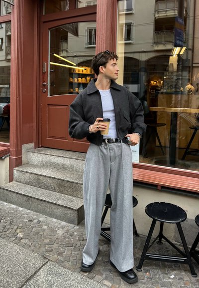 Chaqueta gris corta sobre una blusa gris clara, pantalones grises anchos, zapatos negros. Sosteniendo una taza de café. Escaleras de piedra y taburetes negros visibles.