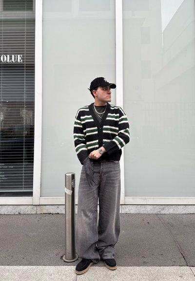 Hombre con cárdigan a rayas, pantalones grises anchos, gorra negra y guantes, de pie en la acera junto a un poste de metal en un entorno urbano.