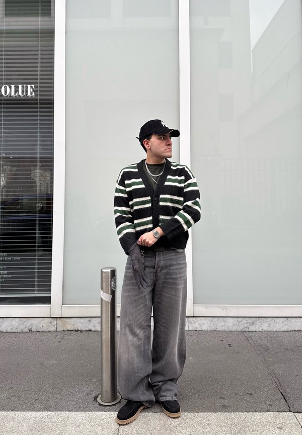 Man draagt een gestreepte cardigan, brede grijze broek, zwarte pet en handschoenen en staat op het trottoir naast een metalen paal in een stedelijke omgeving.
