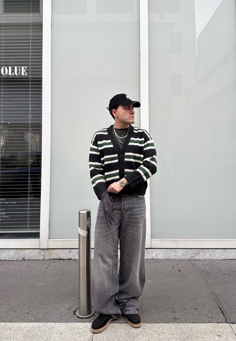 Homme portant un cardigan rayé, un pantalon large gris, une casquette noire et des gants, debout sur le trottoir à côté d'un poteau en métal dans un cadre urbain.