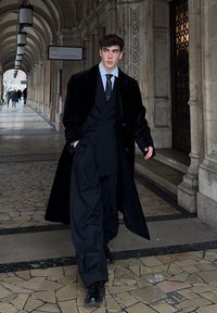 Manteau long en velours noir, costume noir sur mesure avec pantalon ample, chemise blanche, cravate noire, chaussures brillant, installé dans un arc en pierre.