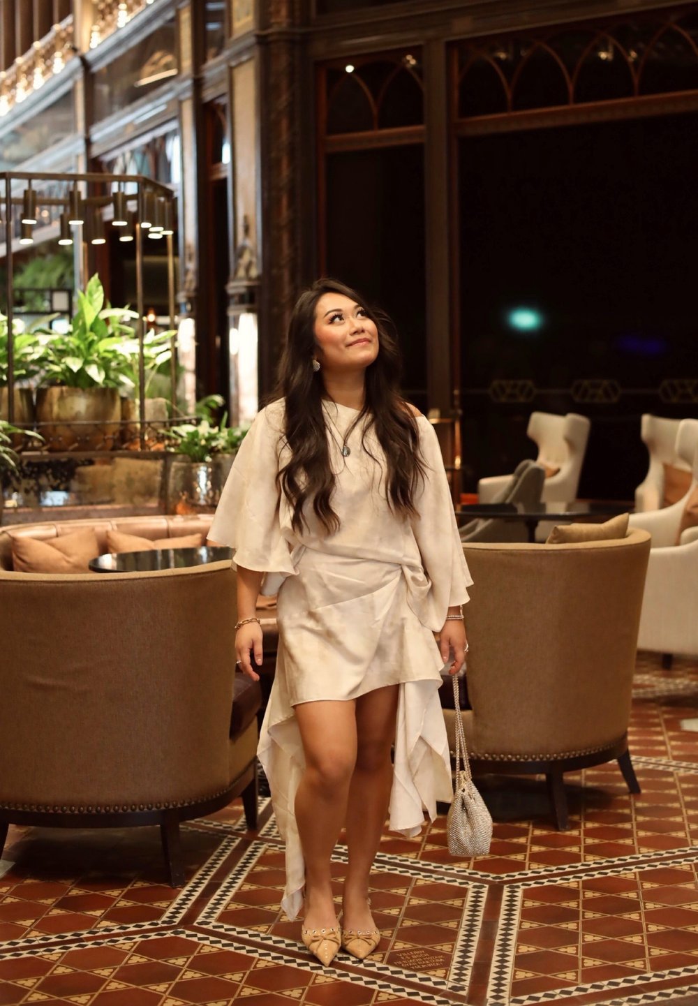 Robe de couleur claire, fluide, avec un design superposé, courte à l'avant et longue à l'arrière. Portée avec des chaussures plates à bout pointu et un petit sac perlé.