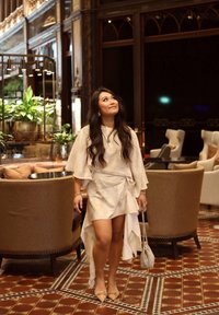 Robe de couleur claire, fluide, avec un design superposé, courte à l'avant et longue à l'arrière. Portée avec des chaussures plates à bout pointu et un petit sac perlé.