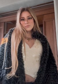 Νεαρή γυναίκα με μακριά ξανθά μαλλιά, φοράει γυαλιά, λευκό τοπ με παγιέτες, στρωμένα χρυσά κολιέ και μαύρο παλτό από ψεύτικη γούνα σε εσωτερικό χώρο.