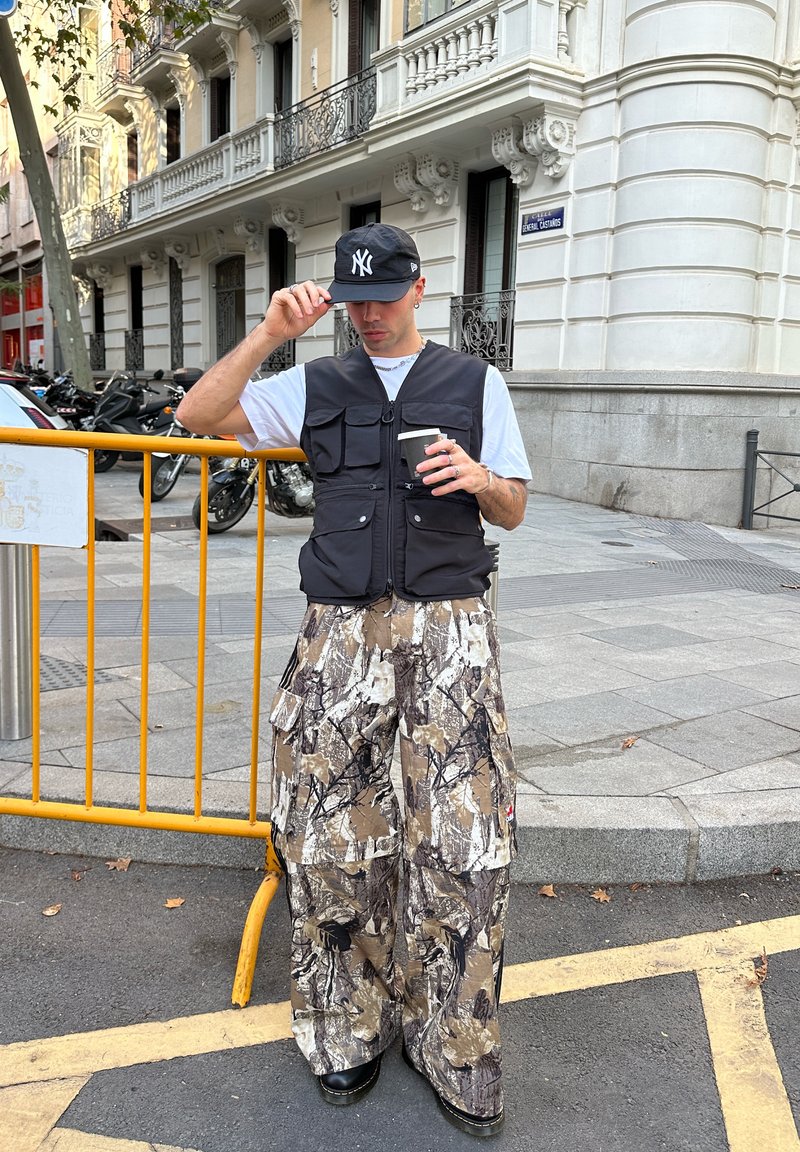 Man in een zwarte pet, zwarte vest en wijde camouflagebroek staat op de stoep van de stad met een koffiekopje bij een gele barrière.