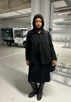 Abrigo de lana negro con botones, combinado con una falda negra y botas de cuero hasta la rodilla. De pie en un área de estacionamiento con un vehículo utilitario detrás.