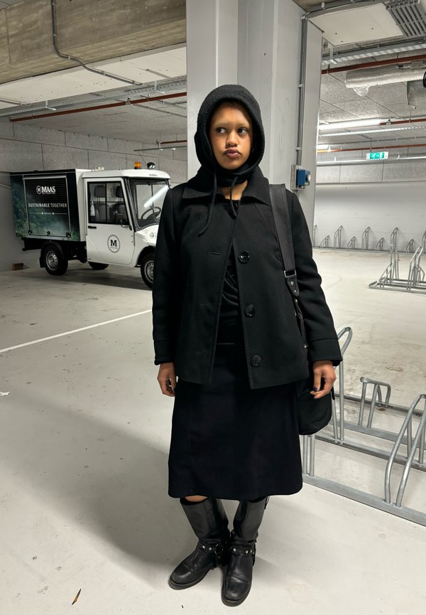 Cappotto di lana nero con bottoni, abbinato a una gonna nera e stivali di pelle al ginocchio. In piedi in un'area di parcheggio con un veicolo utilitario dietro.